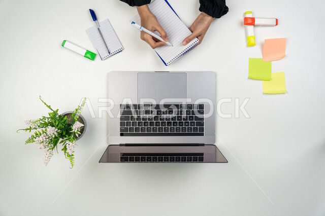 انجاز مهام العمل عن بعد، تسجيل الملاحظات والأفكار، بورتريه علوي عامودي ليدي امرأة عربية خليجية اماراتية تقوم بالكتابة على دفتر وبجانبها أوراق وأقلام ملونة، سيدة سعودية تستخدم جهاز الحاسوب، استخدام الأجهزة التقنية الحديثة في الأعمال المنزلية، خلفية بيضاء