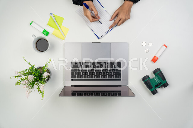 تدوين الملاحظات والأفكار، انجاز مهام العمل عن بعد، بورتريه علوي عامودي ليدي امرأة عربية خليجية اماراتية تقوم بالكتابة على دفتر وبجانبها أوراق وأقلام ملونة ومنظار صغير، سيدة سعودية تستخدم جهاز الحاسوب، استخدام الأجهزة التقنية الحديثة في الأعمال المنزلية، خلفية بيضاء