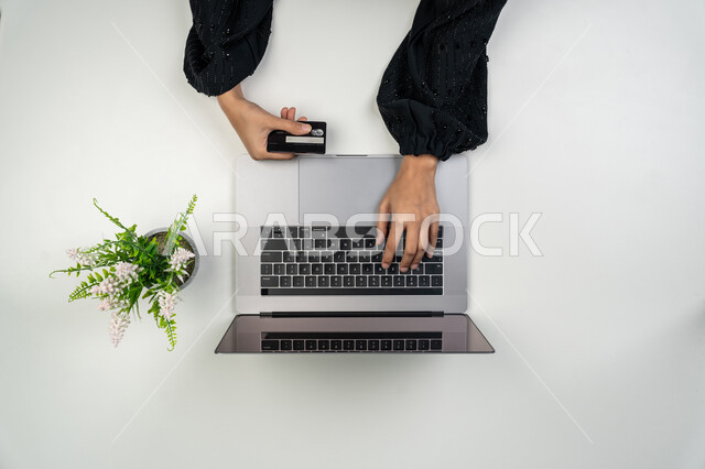 طلب المستلزمات والاحتياجات عن بعد من المنزل، البيع والشراء والتسوق من خلال المواقع الإلكترونية، بورتريه علوي عامودي ليدي إمرأة عربية خليجية اماراتية تقوم باستخدام الحاسوب المحمول، سيدة سعودية تقوم بالدفع أونلاين عبر بطاقة الصراف البنكية، خلفية بيضاء