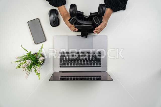 Modern advanced technologies and their integration into work, display of a mobile phone with a blank black screen, use of a laptop in daily life, close-up portrait of the hands of an Arab Gulf Emirati woman holding virtual reality glasses, a Saudi woman with a laptop and a mobile phone on the table next to her, white background