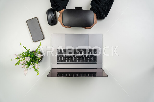 عرض هاتف محمول بشاشة سوداء فارغة، استعمال الحاسوب المحمول في الحياة اليومية, دمج التقنيات الحديثة المتطورة في العمل, بورتريه علوي عامودي لايدي امرأة عربية خليجية اماراتية تمسك نظارة الواقع الافتراضي، سيدة سعودية بجانبها جهاز اللابتوب وموبايل على الطاولة، خلفية بيضاء