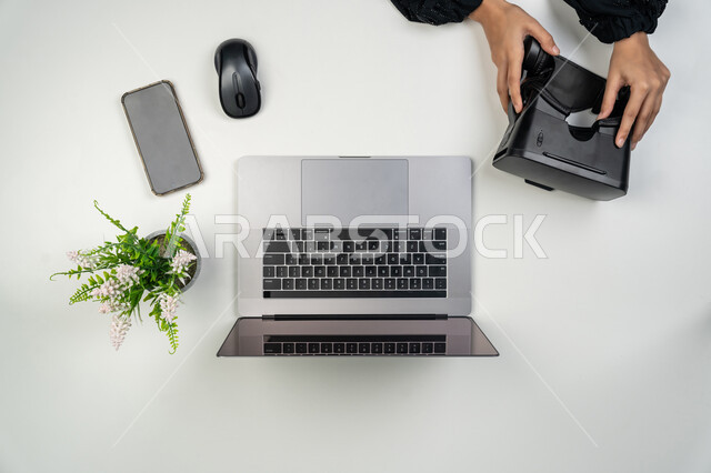استعمال الحاسوب المحمول في الحياة اليومية, دمج التقنيات الحديثة المتطورة في العمل, بورتريه علوي عامودي لايدي امرأة عربية خليجية اماراتية تمسك نظارة الواقع الافتراضي، سيدة سعودية بجانبها جهاز اللابتوب وموبايل على الطاولة، عرض هاتف محمول بشاشة سوداء فارغة، خلفية بيضاء