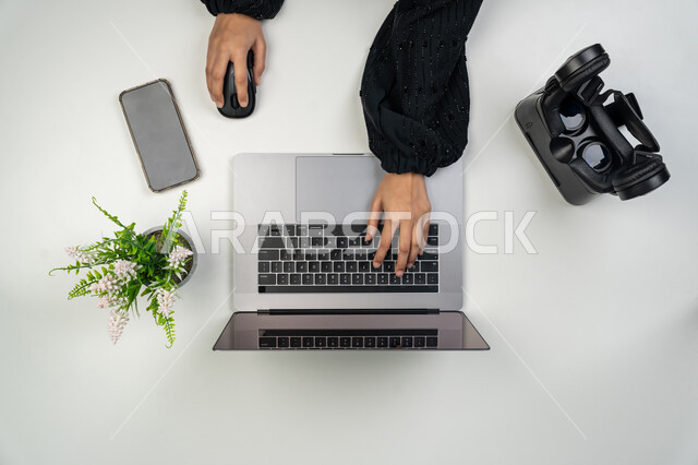 Integrating advanced modern technologies into work, using laptop in daily life, close-up portrait of hands of an Arab Gulf Emirati woman working on laptop, Saudi woman with virtual reality glasses and mobile phone on table, mobile phone display with blank black screen, white background