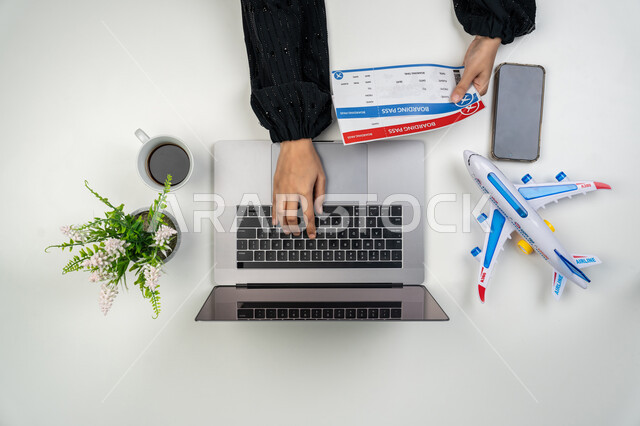 Using websites to search for flights, buying and selling airline tickets online, close-up portrait of an Emirati Gulf Arab woman using a laptop and professional cameras, a young Saudi woman with a passport, a travel ticket, a cup of coffee and a model of an airplane next to her, white background