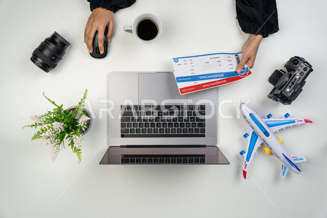 بيع وشراء تذاكر الطيران أونلاين، استخدام المواقع الالكترونية للبحث عن رحلات جوية، بورتريه علوي عامودي ليدي امرأة عربية خليجية اماراتية تستخدم جهاز الحاسوب المحمول كاميرات التصوير الاحترافية، شابة سعودية بجانبها جواز وتذكرة السفر وفنجان قهوة ومجسم لطائرة، خلفية بيضاء