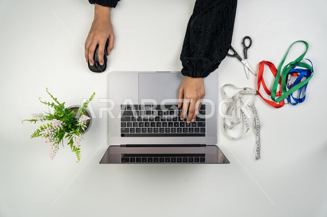 استخدام الأدوات المناسبة لتفصيل الملابس, تصميم ورسم الأزياء, مهن و أعمال نسائية، بورتريه علوي عامودي لأيدي مصممة أزياء عربية خليجية اماراتية تحمل بيدها متر القياس وتقوم بالعمل على الحاسوب المحمول، امرأة سعودية تستخدم التكنولوجيا الحديثة والمتطورة، خلفية بيضاء