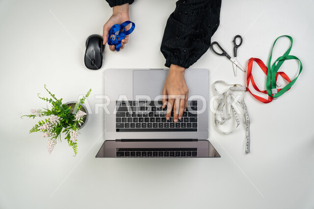 استعمال الأدوات المناسبة لتفصيل الملابس, تصميم ورسم الأزياء, مهن و أعمال نسائية، بورتريه علوي عامودي لأيدي مصممة أزياء عربية خليجية اماراتية تحمل بيدها متر القياس وتقوم بالعمل على الحاسوب المحمول، امرأة سعودية تستخدم التكنولوجيا الحديثة والمتطورة، خلفية بيضاء