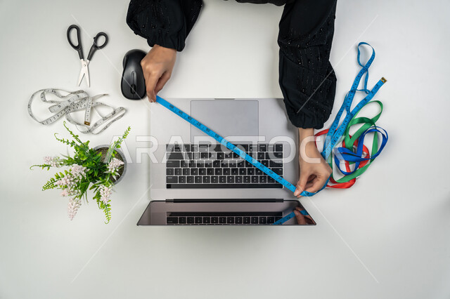 مهن و أعمال نسائية، تصميم ورسم الأزياء, استعمال الأدوات المناسبة لتفصيل الملابس, بورتريه علوي عامودي لأيدي مصممة أزياء عربية خليجية اماراتية تحمل بيدها متر القياس وتقوم بالعمل على الحاسوب المحمول، امرأة سعودية تستخدم التكنولوجيا الحديثة والمتطورة، خلفية بيضاء