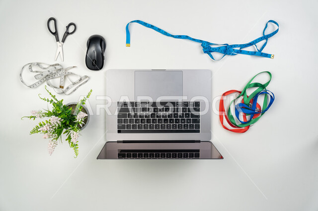 استخدام التكنولوجيا الحديثة والمتطورة، اتقان صنع الثياب، استعمال الأدوات المناسبة لتفصيل الملابس, تصميم ورسم الأزياء, مهن و أعمال سعودية، صورة علوية عامودية لجهاز الحاسوب المحمول مع أمتار القياس والمقص، خلفية بيضاء