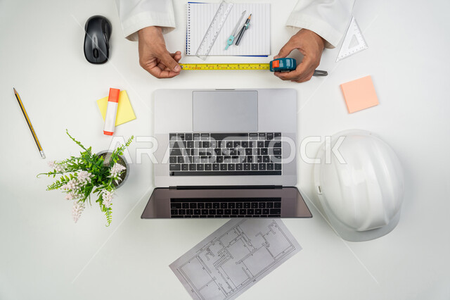 Taking appropriate measurements in metres, using engineering tools and equipment, integrating modern technologies into engineering works, a vertical overhead portrait of the hands of an Arab, Gulf, and Emirati engineer holding the metric tape in his hands, using technology in practical life, white background.