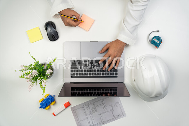 دمج التقنيات الحديثة بالأعمال الهندسية, العمل بالقطاع الهندسي, بورتريه علوي عامودي ليدين مهندس عربي خليجي إماراتي يقوم بالكتابة على لوحة المفاتيح و تدوين الملاحظات على الأوراق الملونة, استخدام التكنولوجيا في الحياة العملية, خلفية بيضاء