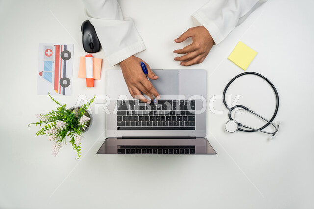 تقديم الخدمات و الاستشارات عن بعد, مفهوم الرعاية الصحية, العمل في القطاع الطبي, بورتريه علوي عامودي ليدين طبيب عربي خليجي إماراتي يقوم بالنقر على لوحة المفاتيح و بجانبه السماعة الطبية, خلفية بيضاء