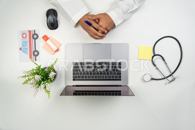 تقديم الخدمات العلاجية عن بعد, العمل في القطاع الطبي, استخدام التقنيات المتطورة في العمل, بورتريه علوي عامودي ليدين طبيب عربي خليجي إماراتي بجانبه السماعة الطبية, خلفية بيضاء