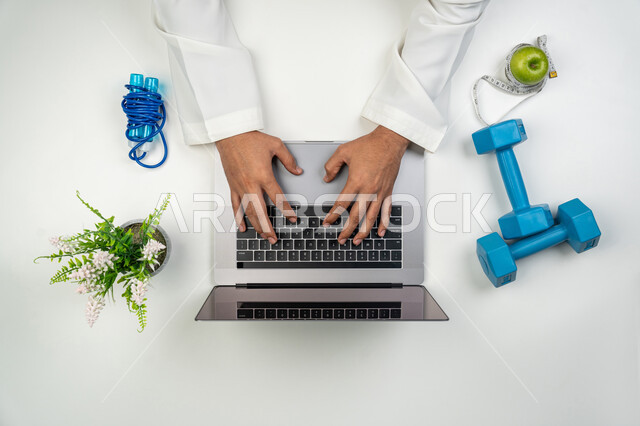 Completing work using modern technical devices, providing physical care services and remote consultations, a vertical overhead portrait of an Arab, Gulf, and Emirati coach writing on a computer, following sports programs via the Internet, white background.