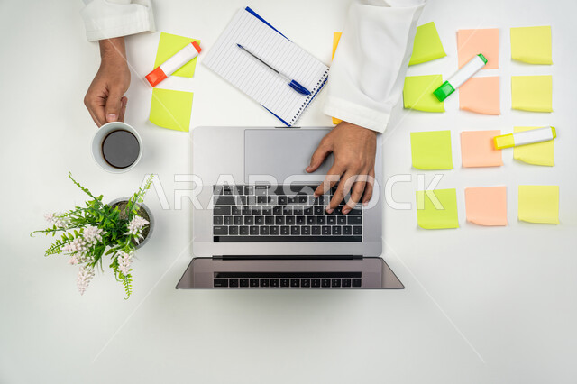 A group of colorful sticky notes on the office table, completing office tasks, using modern technical devices, a vertical overhead portrait of the hands of an Arab, Gulf, Emirati man using a laptop to write reports and send e-mail, white background.