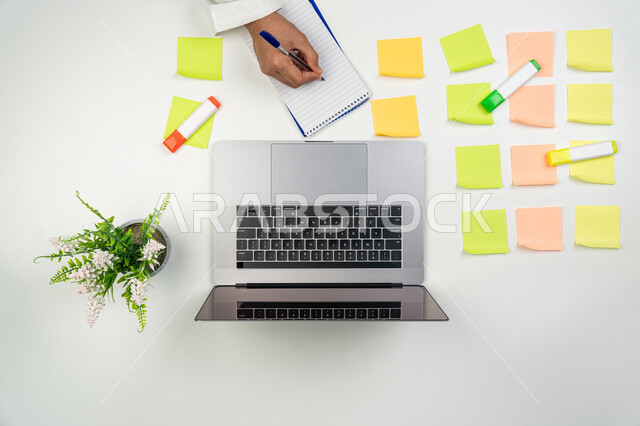 إنجاز المهام المكتبية, استخدام الأجهزة التقنية الحديثة, مجموعة من الملاحظات اللاصقة الملونة على طاولة المكتب, بورتريه علوي عامودي ليدين رجل عربي خليجي إماراتي يقوم بالكتابة على الدفتر, خلفية بيضاء