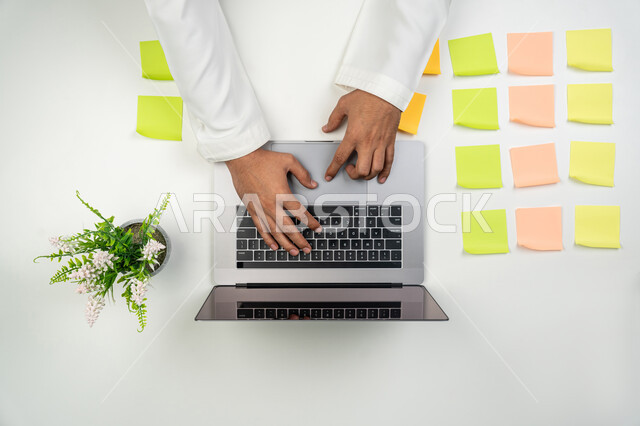 A group of colorful sticky notes on the office table, completing office tasks, using modern technical devices, a vertical overhead portrait of the hands of an Arab, Gulf, Emirati man using a laptop to write reports and send e-mail, white background.