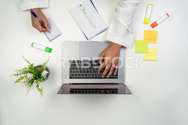 استخدام الأجهزة التقنية الحديثة لإنجاز الأعمال, تسجيل الملاحظات المهمة, مهن و وظائف مكتبية, بورتريه علوي عامودي ليدين رجل عربي خليجي إماراتي يقوم بالنقر على لوحة المفاتيح و الكتابة على الدفتر, خلفية بيضاء