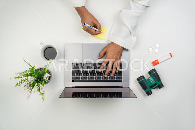 Doing office work, using modern technical devices, comfortable office work environment, vertical overhead portrait of an Arab Gulf Emirati man using a laptop to complete daily tasks and taking notes on colored papers, white background
