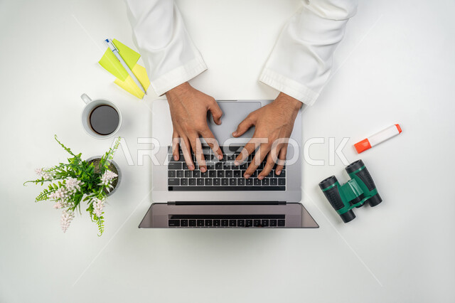 Doing office work, using modern technical devices, a comfortable office work environment, a vertical overhead portrait of the hands of an Arab Gulf Emirati man using a laptop to complete daily tasks, tools that make taking notes happy, white background