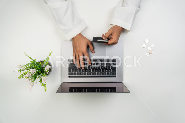 عمليات الشراء أونلاين, الدفع الالكتروني من خلال برامج و تطبيقات اللابتوب, استخدام التقنيات الحديثة و المتطورة, بورتريه علوي عامودي ليدين رجل عربي خليجي إماراتي يمسك بيده بطاقة الائتمان البنكية و يستخدم الحاسوب المحمول, إمكانية تسديد الفواتير عن بعد, خلفية بيضاء