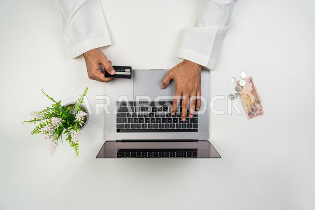 الدفع الالكتروني من خلال برامج و تطبيقات اللابتوب, عمليات الشراء أونلاين, استخدام التقنيات الحديثة و المتطورة, بورتريه علوي عامودي ليدين رجل عربي خليجي إماراتي يمسك بيده بطاقة الائتمان البنكية و يستخدم الحاسوب المحمول, إمكانية تسديد الفواتير عن بعد, خلفية بيضاء
