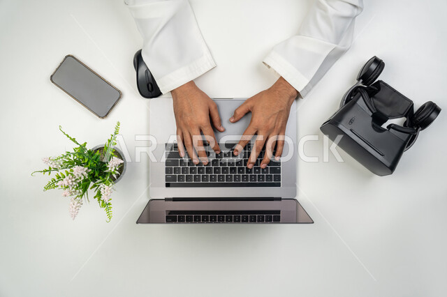 الأجهزة التكنولوجية والتقنيات الحديثة, محاكاة العالم الخيالي, بورتريه علوي عامودي ليدين رجل عربي خليجي إماراتي يقوم بإنجاز العمل على الحاسوب المحمول وبجانبه نظارة الفي ار, خلفية بيضاء