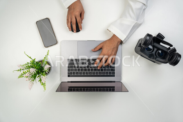 Simulating a virtual world, using modern and advanced technical devices, a vertical overhead portrait of an Arab, Gulf, and Emirati man working on a laptop with VR glasses next to him, a white background.