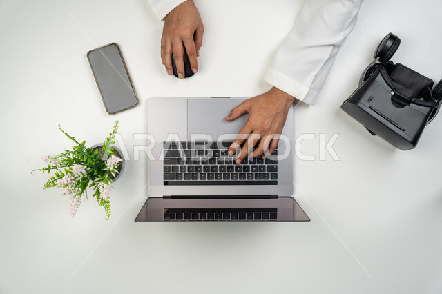 استخدام الأجهزة التقنية الحديثة و المتطورة, محاكاة العالم الافتراضي, بورتريه علوي عامودي ليدين رجل عربي خليجي إماراتي يعمل على الحاسوب المحمول و بجانبه نظارة الفي ار, خلفية بيضاء