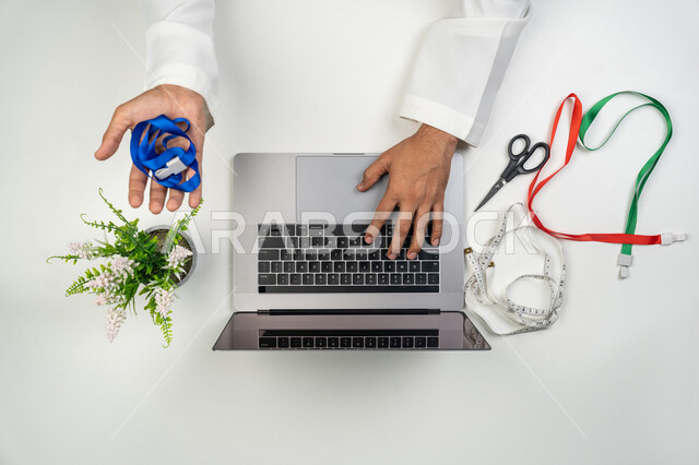 ممارسة مهنة الخياطة, دمج التقنيات الحديثة بالحياة العملية, بورتريه علوي عامودي لمصمم أزياء عربي خليجي إماراتي يمسك بيده شريط أزرق يستخدم جهاز الحاسوب المحمول لإنشاء و تعديل أنماط الخياطة بسرعة و دقة, التواصل مع العملاء و الموردين, الوصول إلى دورات تعليمية عبر الانترنت, خلفية بيضاء