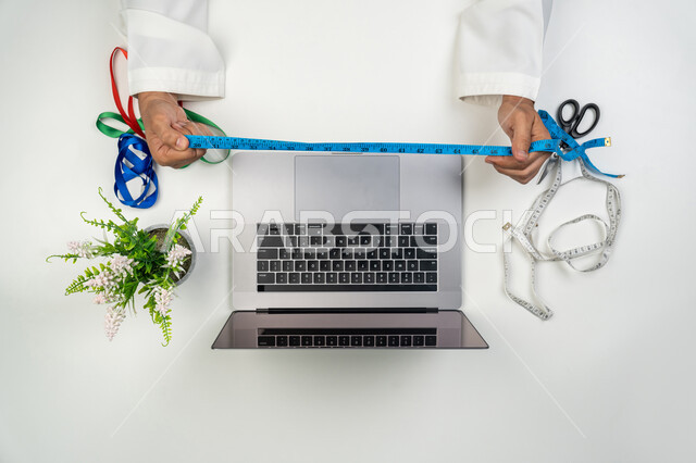 استخدام جهاز الحاسوب الحياة العملية, أعمال و حرف يدوية, ممارسة مهنة الخياطة, بورتريه علوي عامودي لمصمم أزياء عربي خليجي إماراتي يقوم بقياس شيء ما باستخدام الشريط الأزرق, استعمال التقنيات الحديثة, خلفية بيضاء