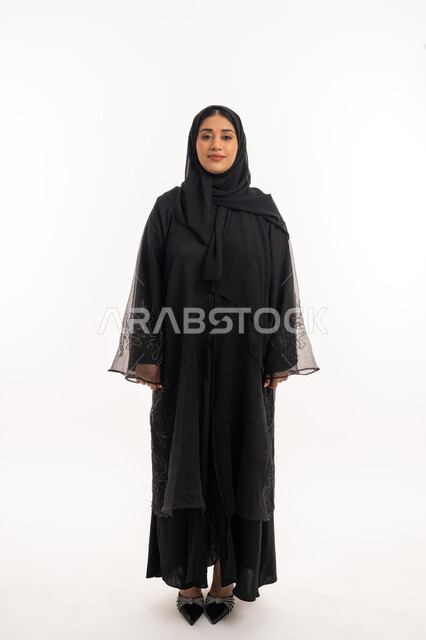 الاهتمام بالمظهر الخارجي، مفهوم الانوثة والاناقة، ايماءات السرور والسعادة، بورتريه لامرأه عربيه خليجيه اماراتيه مبتسمة ترتدي عباءة سوداء تنظر إلى الكاميرا بتعابير الفرح، شابة سعودية انيقة، صورة الجسم بالطول الكامل، خلفية بيضاء