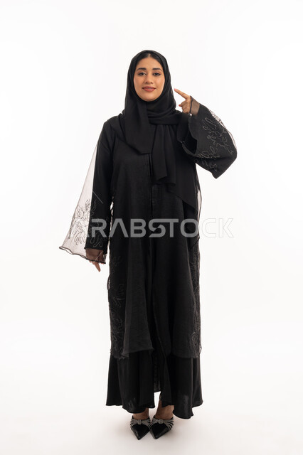 إشارات وحركات يدين تدل على هذا المكان (هنا)، تحديد موقع شيء معين، بورتريه لامرأة عربية خليجية اماراتية محجبة ترتدي عباءة سوداء تشير بإصبع السبابة إلى وجهها، شابة سعودية انيقة، صورة الجسم بالكامل، خلفية بيضاء