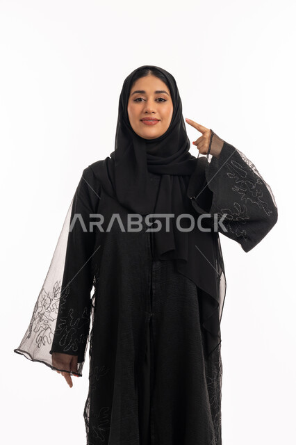 شابة سعودية انيقة، تحديد موقع شيء معين، إشارات وحركات يدين تدل على هذا المكان (هنا)، بورتريه لامرأة عربية خليجية اماراتية محجبة ترتدي عباءة سوداء تشير بإصبع السبابة إلى وجهها، خلفية بيضاء