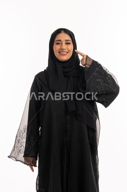 تحديد موقع شيء معين، إشارات وحركات يدين تدل على هذا المكان (هنا)، بورتريه لامرأة عربية خليجية اماراتية محجبة ترتدي عباءة سوداء تشير بإصبع السبابة إلى وجهها، شابة سعودية انيقة، خلفية بيضاء