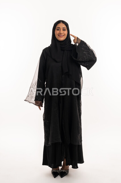 تحديد موقع شيء معين، إشارات وحركات يدين تدل على هذا المكان (هنا)، بورتريه لامرأة عربية خليجية اماراتية محجبة ترتدي عباءة سوداء تشير بإصبع السبابة إلى وجهها، شابة سعودية انيقة، صورة الجسم بالكامل، خلفية بيضاء