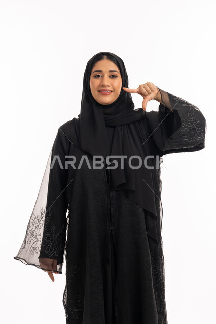 شابة سعودية انيقة، تحديد موقع شيء معين، إشارات وحركات يدين تدل على هذا المكان (هنا)، بورتريه لامرأة عربية خليجية اماراتية محجبة ترتدي عباءة سوداء تشير بإصبع السبابة إلى وجهها، خلفية بيضاء