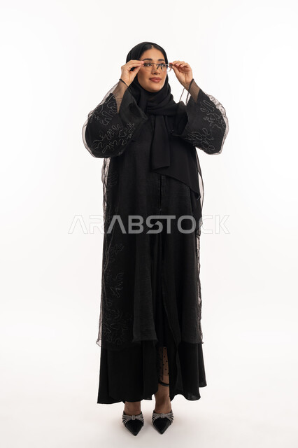 الاناقة والاهتمام بالمظهر الخارجي، بورتريه لامرأة عربية خليجية اماراتية محجبة ترتدي عباءة سوداء تمسك بيدها نظارة طبية، ارتداء النظارات الخاصة بضعف النظر، تحسين الرؤية ومحاولة تعديل البصر، شابة سعودية انيقة، الجسم بالطول الكامل، خلفية بيضاء