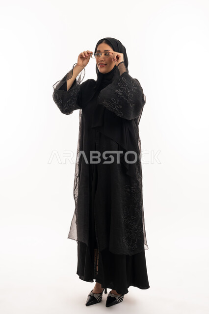 تحسين الرؤية ومحاولة تعديل البصر، ارتداء النظارات الخاصة بضعف النظر، الاناقة والاهتمام بالمظهر الخارجي، بورتريه لامرأة عربية خليجية اماراتية محجبة ترتدي عباءة سوداء تمسك بيدها نظارة طبية، شابة سعودية انيقة، الجسم بالطول الكامل، خلفية بيضاء