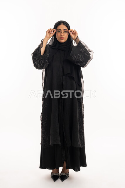 ارتداء النظارات الخاصة بضعف النظر، الاناقة والاهتمام بالمظهر الخارجي، بورتريه لامرأة عربية خليجية اماراتية محجبة ترتدي عباءة سوداء تمسك بيدها نظارة طبية، تحسين الرؤية ومحاولة تعديل البصر، شابة سعودية انيقة، الجسم بالطول الكامل، خلفية بيضاء