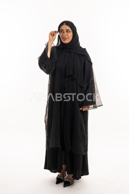 الاناقة والاهتمام بالمظهر الخارجي، بورتريه لامرأة عربية خليجية اماراتية محجبة ترتدي عباءة سوداء تمسك بيدها نظارة طبية، ارتداء النظارات الخاصة بضعف النظر، تحسين الرؤية ومحاولة تعديل البصر، شابة سعودية انيقة، الجسم بالطول الكامل، خلفية بيضاء