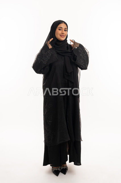 تحديد موقع شيء معين، إشارات وحركات يدين تدل على هذا المكان (هنا)، بورتريه لامرأة عربية خليجية اماراتية محجبة ترتدي عباءة سوداء تشير بإصبعي السبابة إلى وجهها، شابة سعودية انيقة، صورة الجسم بالكامل، خلفية بيضاء