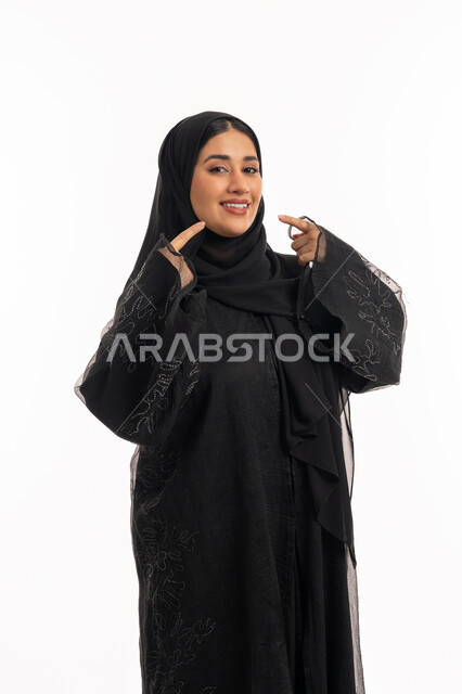 إشارات وحركات يدين تدل على هذا المكان (هنا)، تحديد موقع شيء معين، بورتريه لامرأة عربية خليجية اماراتية محجبة ترتدي عباءة سوداء تشير بإصبع السبابة إلى وجهها، شابة سعودية انيقة، خلفية بيضاء