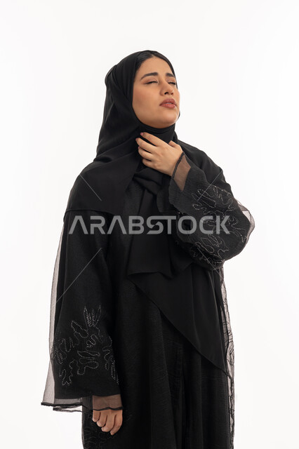 Feeling sick and coughing, expressions of pain and fatigue, influenza and pneumonia, portrait of a veiled Arab Gulf Emirati woman wearing a black abaya, putting her hand to her throat in gestures of pain, a sick Saudi young woman, white background