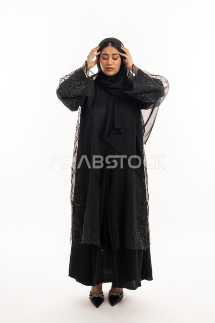 Suffering from migraine pain, feeling headache and migraine, need for calm and rest, portrait of a veiled Gulf Arab Emirati woman wearing a black abaya, putting her hands on her head with gestures of fatigue and exhaustion, a sick Saudi young woman, full body image, white background