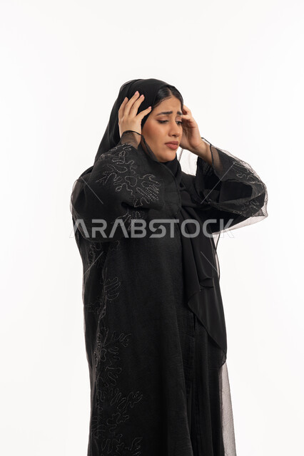 Need for peace and comfort, feeling headache, suffering from migraine pain, side portrait of a veiled Gulf Arab Emirati woman wearing a black abaya, putting her hands on her head with gestures of fatigue and exhaustion, a sick Saudi young woman, white background