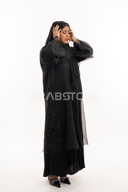 الحاجة للهدوء والراحة، الإحساس بألم الرأس والصداع، المعاناة من آلام الشقيقة، بورتريه من الجانب لإمرأة عربية خليجية اماراتية محجبة ترتدي عباءة سوداء تضع يديها على رأسها بايماءات التعب والارهاق، شابة سعودية مريضة، صورة الجسم بالطول الكامل، خلفية بيضاء
