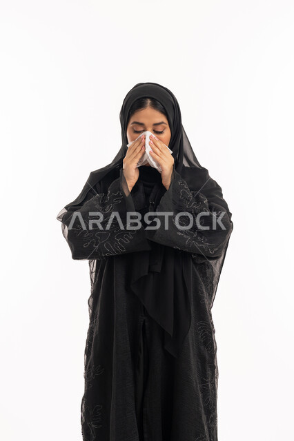 Feeling tired and sick, using tissues while sneezing, infection with the Corona virus Covid19, portrait of a veiled Arab Gulf Emirati woman wearing a black abaya, facial and hand gestures indicating sneezing, a sick young Saudi woman, white background