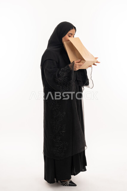 إيماءات التقيؤ والغثيان، الإحساس بالمغص والوجع، الحاجه للمساعدة وطلب العلاج، بورتريه  من الجانب لامرأة عربية خليجية إماراتية ترتدي عباءة سوداء تحمل بيدها كيس ورقي، شابة سعودية مريضة، صورة لكامل الجسم، خلفية بيضاء