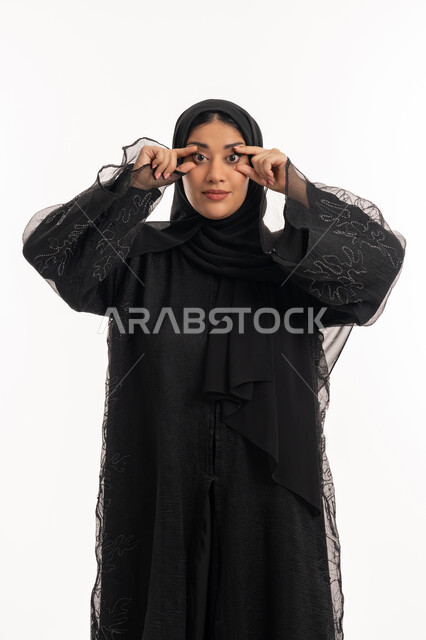 التركيز بالنظر على شيء معين، المعاناة من ضعف البصر، محاولة الرؤية بوضوح، بورتريه لامرأة عربية خليجية اماراتية محجبة ترتدي عباءة سوداء يرفع يديها على شكل نظارات بجانب عينيها، شابة سعودية انيقة، خلفية بيضاء